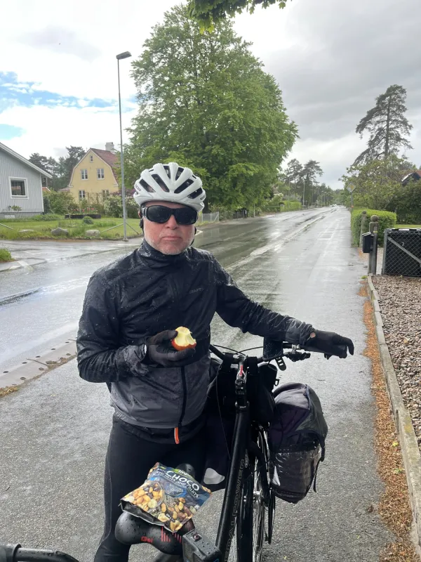 Oskar Jonsson äter äpple och står vid tandemcykeln intill vägen, i regnet.