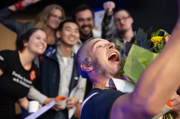 Glad man tar en selfie tillsammans med fem personer i bakgrunden vars ansikten är lite suddiga.