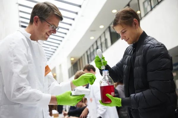 Forskare i vit rock och gröns plasthandskar står bredvid elev med gröna plasthandskar. Eleven håller i en flaska med röd vätska.