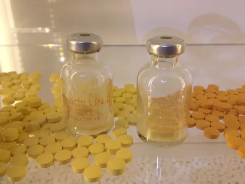 Two pill jars made of glass with the word penicillin on it, surrounded by yellow pills on a table