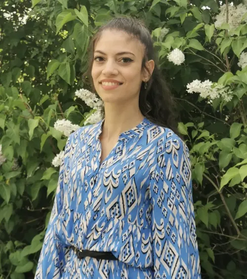 Portrait of a woman, standing in front of a bush with green leaves and white blossoms.