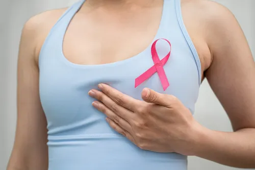 Woman in a light blue tank top with a pink ribbon holds her hand under one of her breasts.