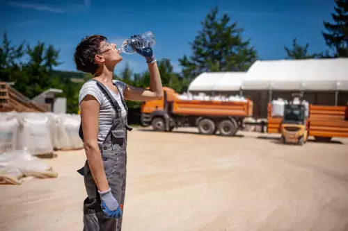 Young woman in work wear drinks water from a bottle on a dusty road in front of lorries