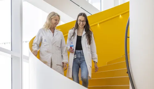 Two women in lab coats walking down yellow stairs.