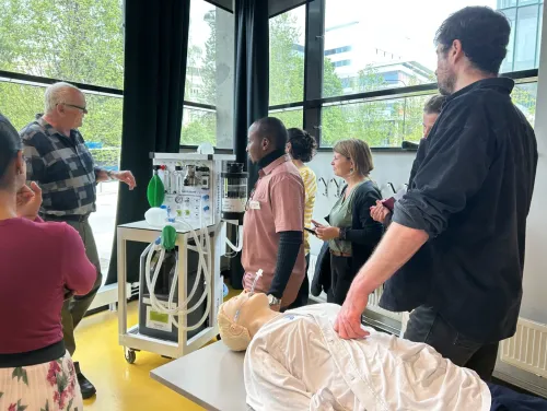 A group of people are standing around a machine, and a first aid manikin is lying on a table
