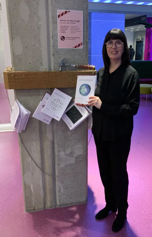 Woman standing with her thesis in hand in front of a pillar where several theses are nailed.