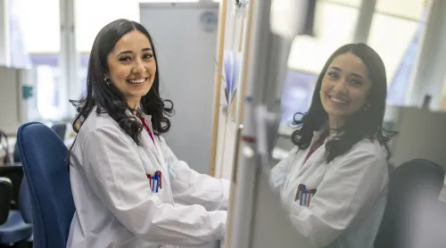 Woman in lab coat looks into the camera and smiles. Her face is reflected in a glass door.