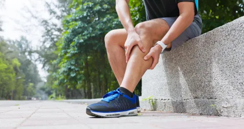 Closeup young man is sporting in park he stops and sits down by roadside chair suffers from calf cramping with hands touching massage the painful point straightening legs pull foot for ease