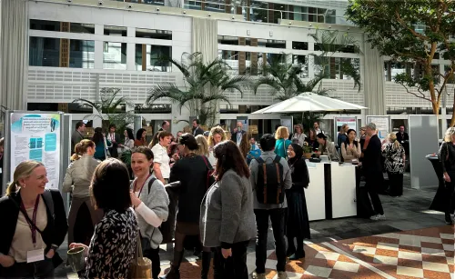 People talking to each other in a bright room, with big roof top windows. The sun i shining on big indoor palm trees.