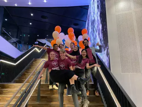Happy students stnading in a stair. They are wearing purple t-shirts with KI logo. Orange and white ballons are visible in the background.