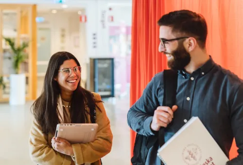 Two students laughing on campus. One of them is holding a laptop, the other a notebook with the logo of Karolinska Institutet.