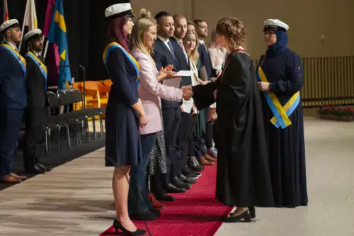 Students on stage receive envelopes with a greeting from the President, which in this photo is handed out by the Academic vice president of higher education wearing an academic gown.