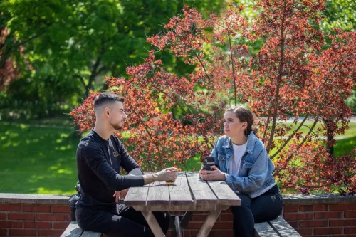 Two students sit at a wooden table at Jöns Jacob&#039;s outdoor terrace at Solna campus on a sunny spring day.