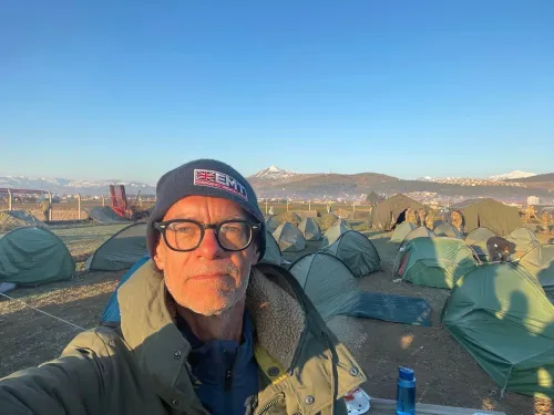 Johan von Schreeb in front of rows of green tents set up on dry ground with snow-capped mountains in the background under a clear blue sky.