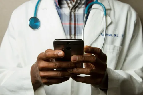 A person in a doctor&#039;s coat with a stethoscope around his neck is holding a smartphone.