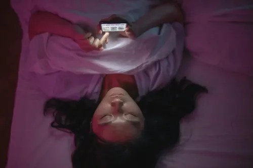 Young woman lying on her back in a bed lookaing at her phone. The light is dimmed.