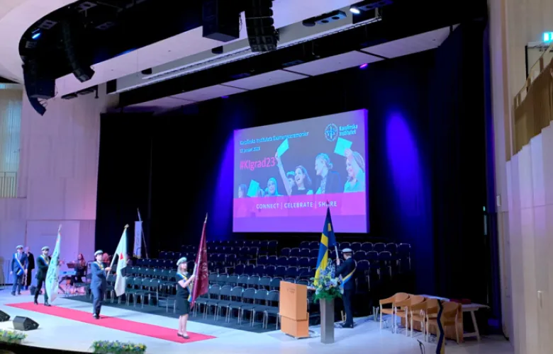 The student ushers are carrying the flags up on stage in Erling Persson-hall. On big screen there is a photo from a previous graduation ceremony with information about what # to use on social media.